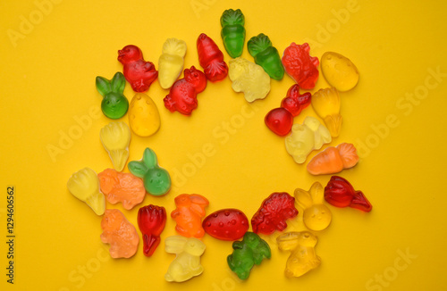 A festive wreath made of colorful gummy candies shaped like Easter bunnies, chicks, and carrots on a yellow background. A playful and bright holiday composition.