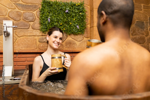 A multi-ethnic couple in their 20s and 30s enjoy a day at a spa with beer tubs.The woman looks happily at the boy with a mug of beer.Beer spa concept.