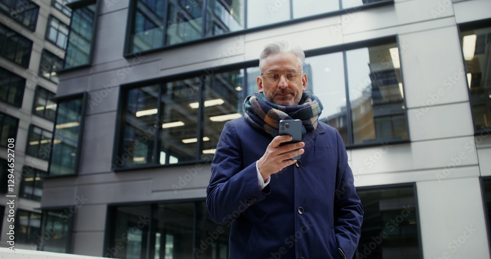 A gray-haired man with a scarf around his neck uses a mobile phone while standing outdoors in the business center of the city among glass skyscrapers. Dynamic circular video. Video in 4k, red komodo A gray-haired man with a scarf around his neck uses a mobile phone while standing outdoors in the business center of the city among glass skyscrapers. Dynamic circular video. Video in 4k, red komodo