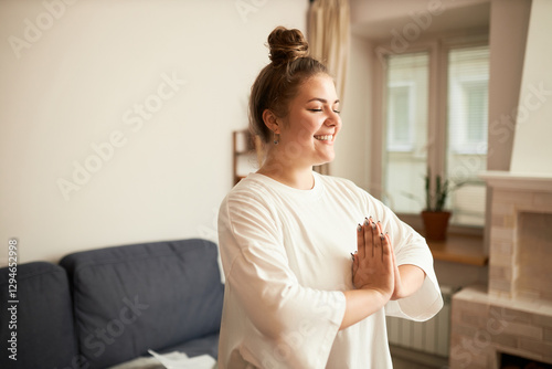 Side view of young smiling overweight female meditating in home interior, visualizing her better version of herself , standing with closed eyes and keeping hands in prayer position