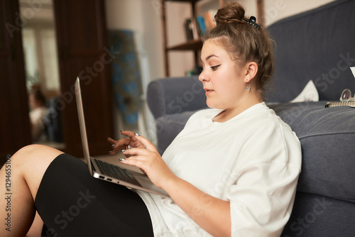 Side view of female interpreter freelance worker in home clothes sitting on floor next to sofa with laptop on knees, typing while searching for right equivalent word online involved in working process