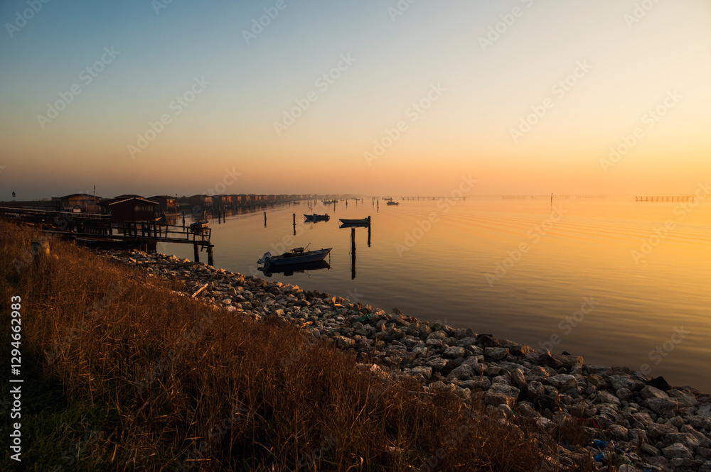 Obraz premium winter sunset over the Scardovari lagoon with fishing boats and cabins inside the delta of the river Po, Porto Tolle, Veneto