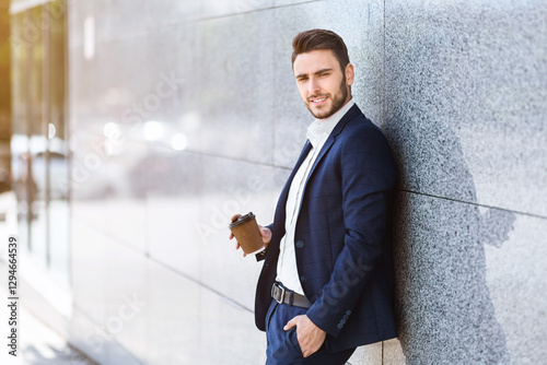 Handsome business manager with takeaway coffee leaning to office building wall in modern megapolis