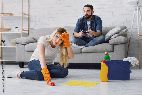 Cleaning, duties and sexism at home. Man has fun with video games, tired wife clings to her head and do housekeeping