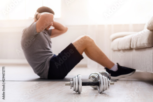 Guy does exercises for abs, shove his legs under couch in living room in daylight, at interior. Dumbbells on floor, close up