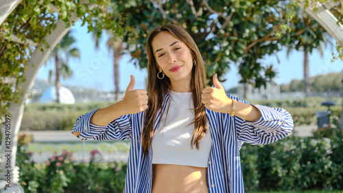 A young businesswoman in a tree-filled park on a spring day, looking at the camera while giving a thumbs-up gesture as a sign of approval