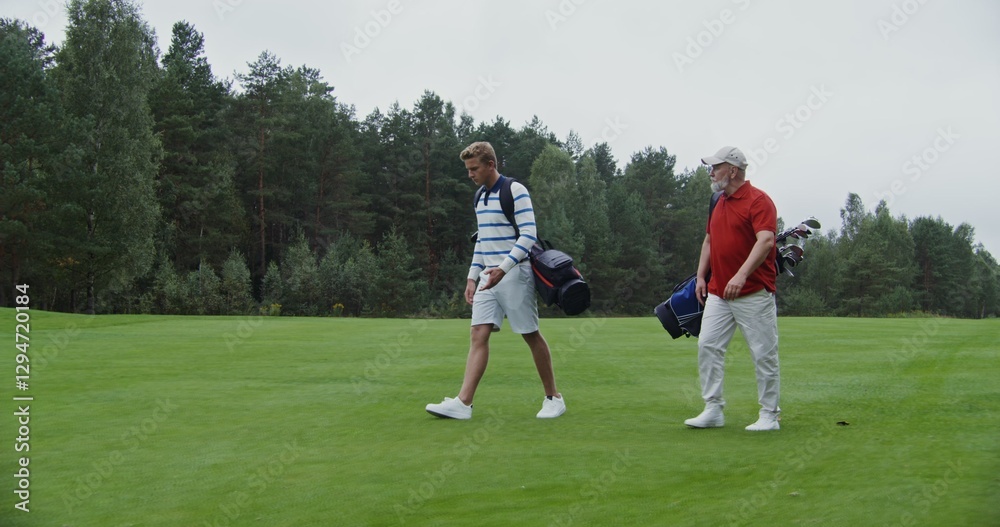 Two men of European appearance with a large age difference walk the golf course with bags of clubs and equipment, talking to each other. Preparing to play golf on a large, well-groomed golf course Two men of European appearance with a large age difference walk the golf course with bags of clubs and equipment, talking to each other. Preparing to play golf on a large, well-groomed golf course