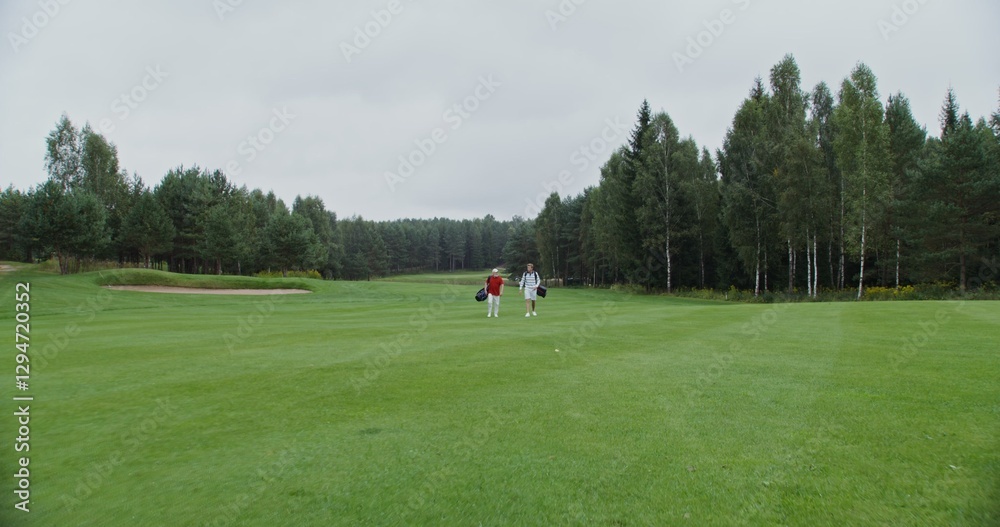 father and son walk along the golf course, carrying golf clubs in their bags. Two men of different ages are going to play golf on a large well-groomed field surrounded by dense green forest father and son walk along the golf course, carrying golf clubs in their bags. Two men of different ages are going to play golf on a large well-groomed field surrounded by dense green forest