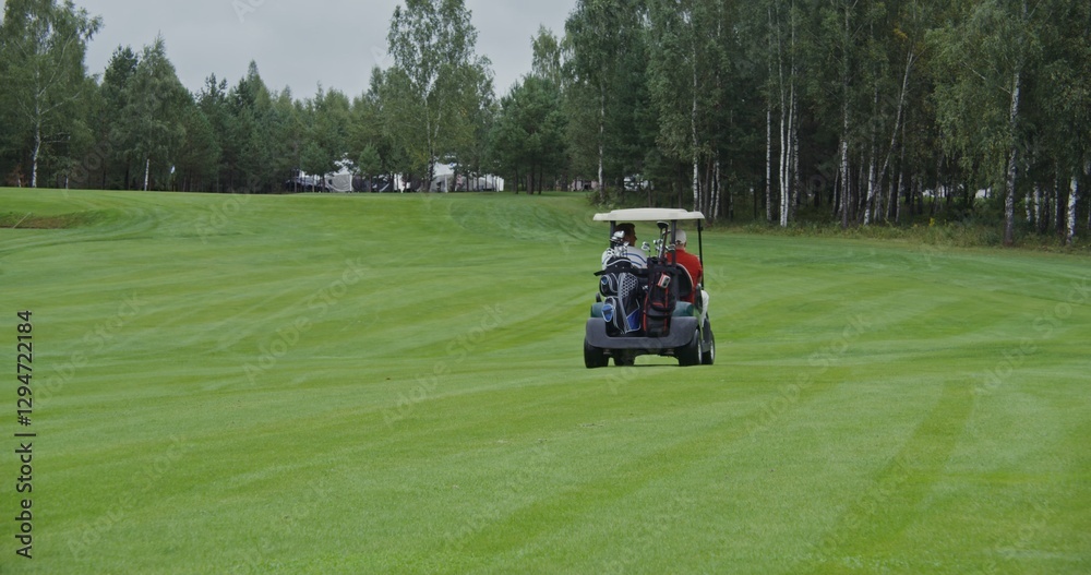 Two men of European appearance are driving a golf car along the golf course, carrying bags of golf equipment and talking to each other on a rainy day, shot from behind, a view of the car receding Two men of European appearance are driving a golf car along the golf course, carrying bags of golf equipment and talking to each other on a rainy day, shot from behind, a view of the car receding