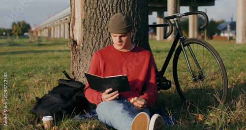 A young man of European appearance in a knitted hat reads a book and eats a sandwich, sitting on the ground under a tree next to the bridge. The bike stands under the tree next to the guy