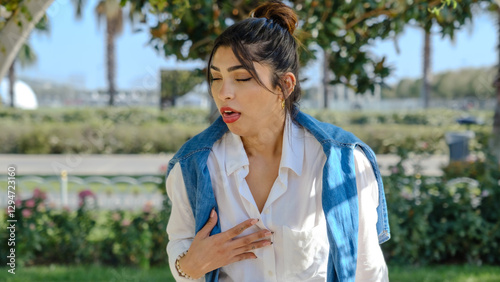 A young brunette businesswoman in a tree-filled park on a spring day, looking at the camera with facial expressions that indicate she is feeling nauseous