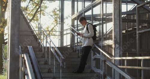 A young man of European appearance in a knitted hat uses a mobile phone, and drinks coffee while standing on the steps of a metal staircase.