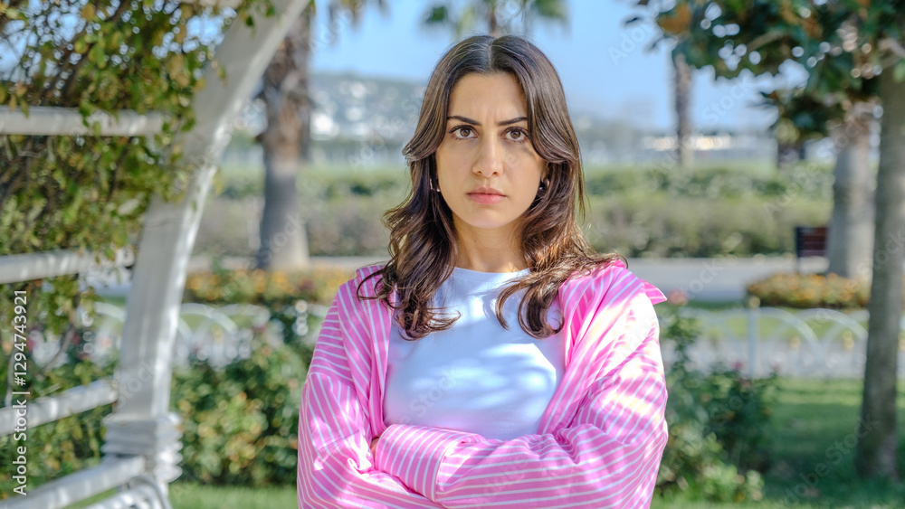 A young brunette businesswoman in a tree-filled park on a spring day, looking at the camera with facial expressions that show she is feeling bored