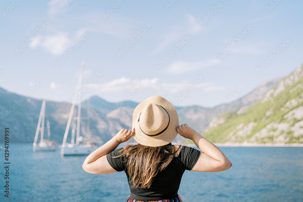 Fototapeta premium Woman stands on the shore holding a straw hat on her head and looks at the sailboats. Back view