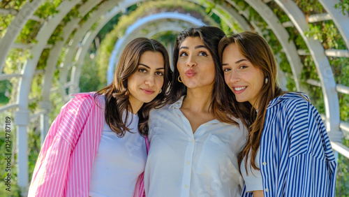 Three young female friends in a tree-filled park on a spring day, looking at the camera while blowing kisses