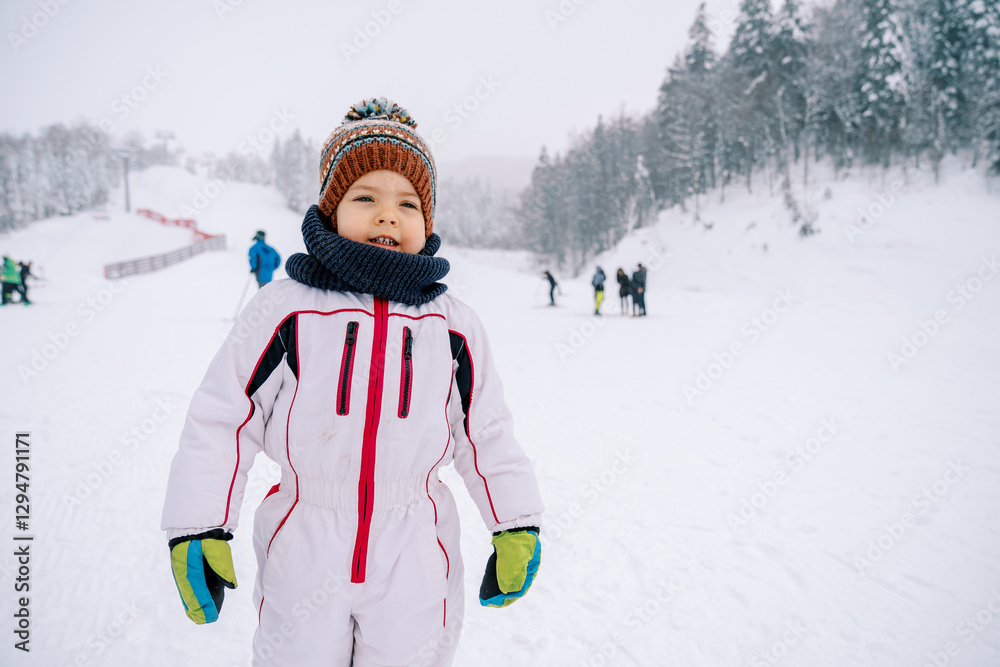 Little smiling girl in a ski suit stands on a ski slope Little smiling girl in a ski suit stands on a ski slope