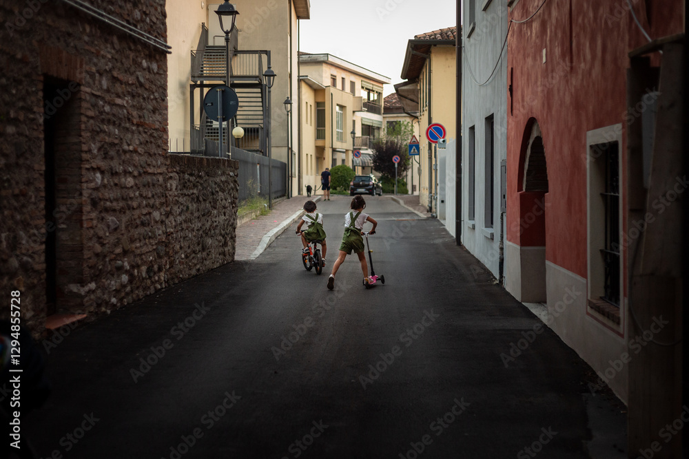 rear view of two small girls riding bicycle and scooter in the street of a town rear view of two small girls riding bicycle and scooter in the street of a town