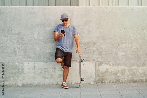 Young skateboarder using smartphone leaning against concrete wall