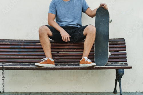 Unrecognizable skater resting on bench holding skateboard after exercising