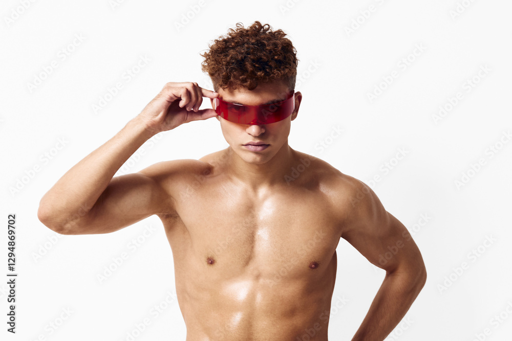 Fototapeta premium Confident young man with curly hair wearing stylish red sunglasses, showcasing a strong physique against a clean white background. Fashion and confidence concept.