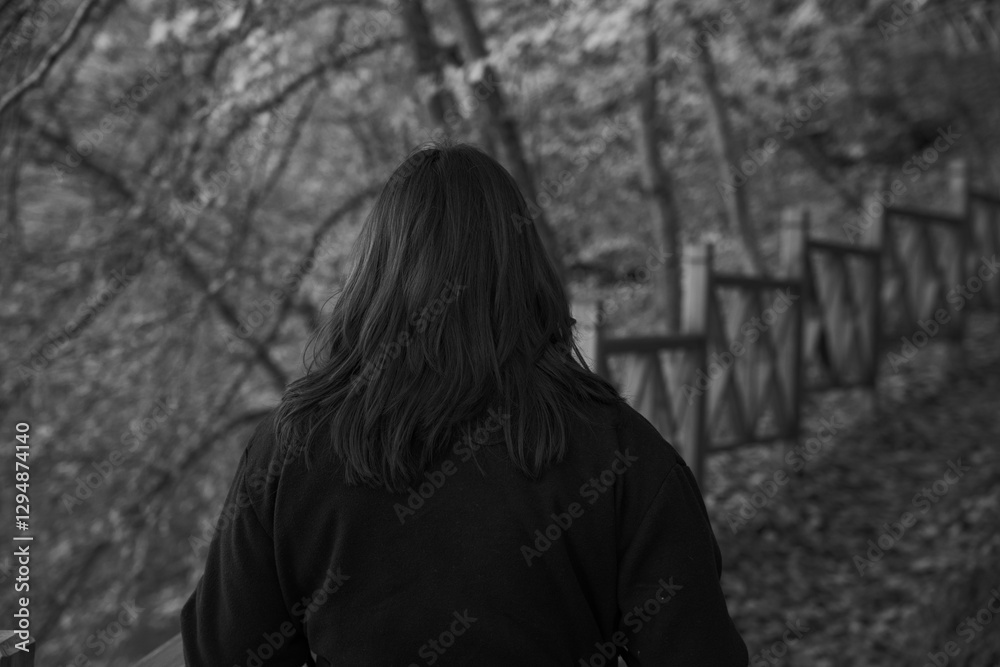 Woman walking lonely on serene forest path with wooden fence in black and white. Melancholic autumn stroll. Deep in thought. Back view Woman walking lonely on serene forest path with wooden fence in black and white. Melancholic autumn stroll. Deep in thought. Back view