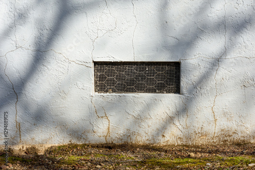 Rectangular crawl space vent, ventilation opening with a metal mesh cover. White aging stucco wall, surrounded by cracks and weathering