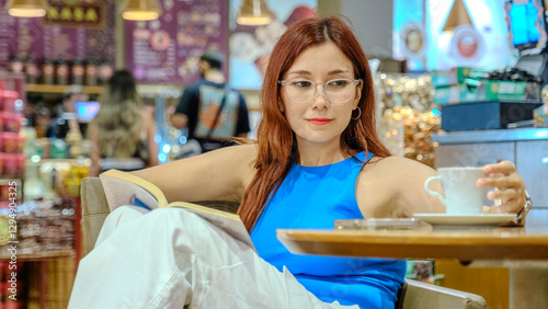 A beautiful young woman with red hair and glasses, wearing a blue t-shirt, is sitting in a lovely café on a summer evening, enjoying reading a book