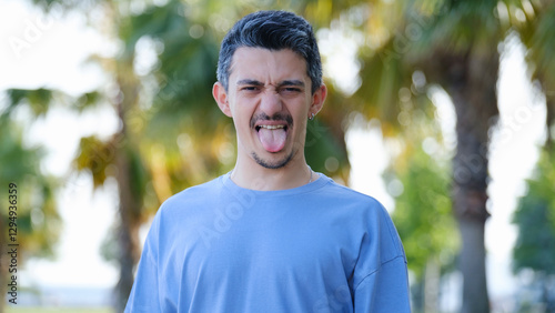 A dark-haired young man in a tree-filled park, looking at the camera and sticking out his tongue while having fun
