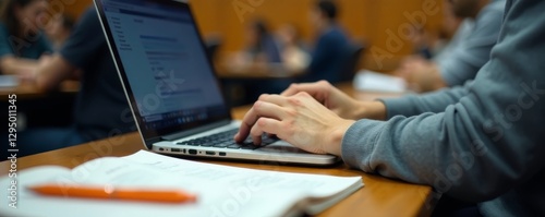 Student's hands typing on laptop, notes and pen visible on desk in lecture hall, remote learning, hybrid learning, college