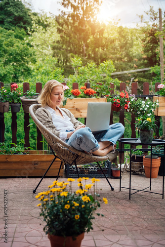 Young adult Caucasian blonde woman sitting on cottage terrace using laptop, online work in the countryside concept.  Intelligent attractive female successful freelancer does business in Her own home photo