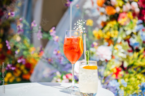 Glass of Aperol spritz cocktail, decorated with slices of orange and flower branch next to the other cocktail with a slice of orange, rosemary and ice on background of sunny flowers