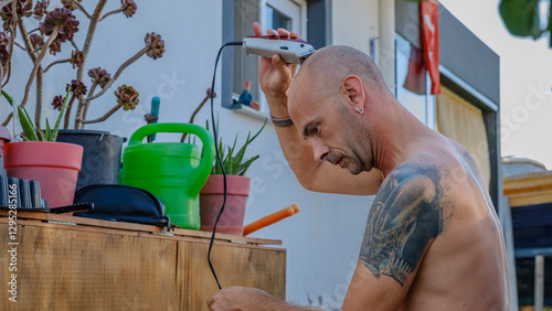 A handsome middle-aged man shaving his hair and beard topless in the tree-filled garden of his house on a sunny summer day