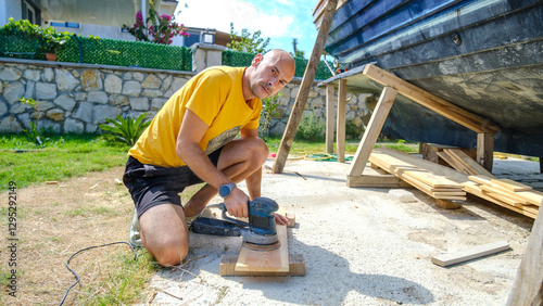A middle-aged man sanding timber with a sander in the garden of his house on a summer day