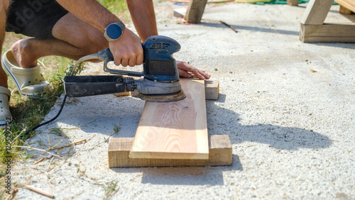 A middle-aged man sanding timber with a sander in the garden of his house on a summer day