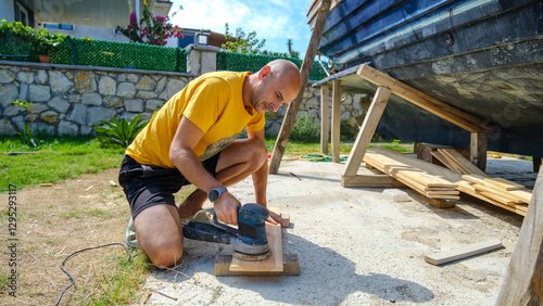 A middle-aged man sanding timber with a sander in the garden of his house on a summer day