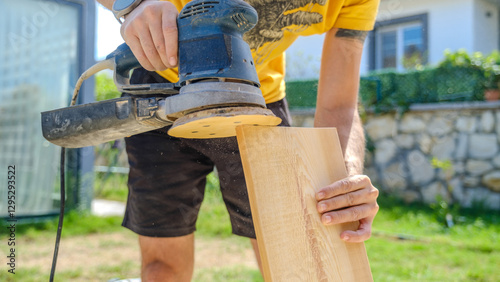 A middle-aged man sanding timber with a sander in the garden of his house on a summer day