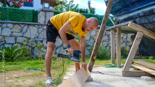 A middle-aged man sanding timber with a sander in the garden of his house on a summer day