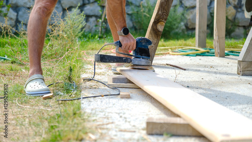 A middle-aged man sanding timber with a sander in the garden of his house on a summer day