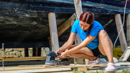 A young woman is sanding timber with a sander in the garden of her home on a summer day.