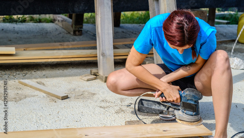 A young woman is sanding timber with a sander in the garden of her home on a summer day.