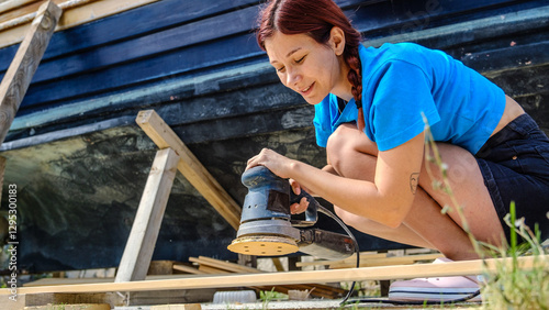 A young woman is sanding timber with a sander in the garden of her home on a summer day.