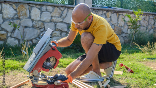A middle-aged man cutting timber with a circular saw in the garden of his house on a summer day