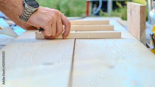 A middle-aged man measures with a tape measure to make repairs with lumber in his garden on a summer day.