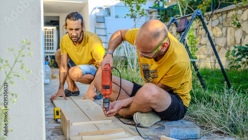 Two middle-aged men using cordless screwdrivers to drive screws while making furniture with timber in the garden of their house on a summer day