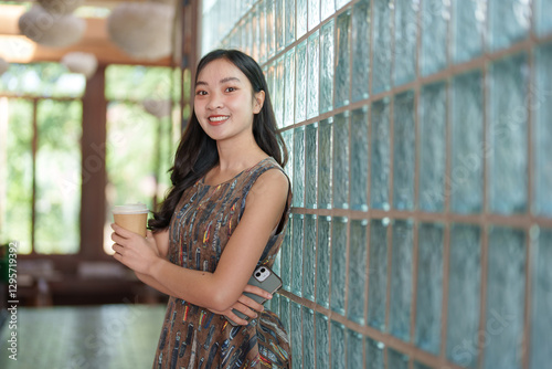 Cheerful young asian woman in a patterned dress enjoys her coffee break, holding a takeaway cup and her smartphone while leaning against a stylish glass brick wall Free Download Cheerful young asian woman in a patterned dress enjoys her coffee break, holding a takeaway cup and her smartphone while leaning against a stylish glass brick wall - GFXLoad