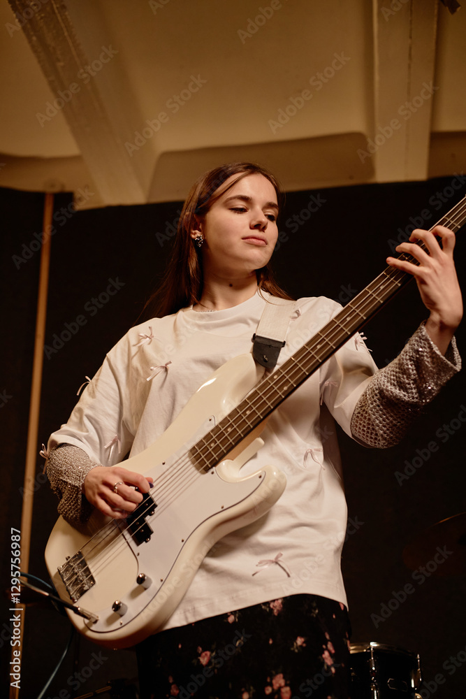 Obraz premium Vertical shot of confident teenage female artist hitting chord with deft fingers on electric bass guitar while playing punk rock music in recording studio