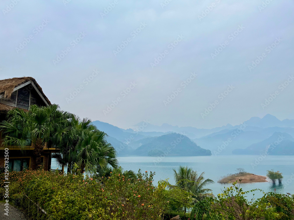Scenic view of a lake surrounded by mountains, lush greenery, and a quaint thatched-roof building under a slightly overcast sky. Scenic view of a lake surrounded by mountains, lush greenery, and a quaint thatched-roof building under a slightly overcast sky.
