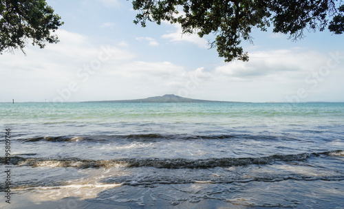 Gentle waves lapping on beach. Rangitoto Island framed by Pohutukawa trees. Takapuna Beach. Auckland.