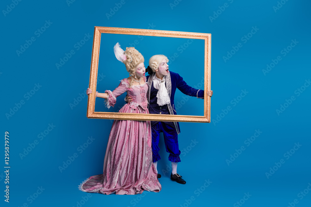 Fototapeta premium Young couple dressed in extravagant baroque costumes holding a golden frame against a vibrant blue background