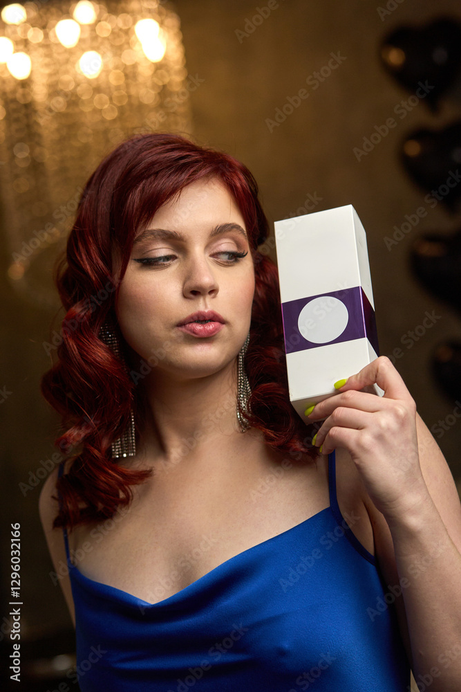 Elegant redhead woman in blue dress holding a white box with purple label and thoughtful expression, glamour concept